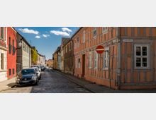 Eine gepflasterte Straße mit parkenden Autos, flankiert von historischen Häusern in verschiedenen Farben, darunter ein orangefarbenes Fachwerkhaus mit weißen Fenstern. Im Vordergrund steht ein Verkehrsschild für die Einfahrtverbot, und blauer Himmel mit wenigen Wolken bildet den Hintergrund.