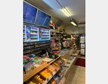 Ein kleines Ladengeschäft mit einer Verkaufstheke, auf der Süßigkeiten, Feuerzeuge und kleine Artikel ausgestellt sind. Im Hintergrund befinden sich Regale mit Tabakwaren, Getränken, Zeitschriften und Zeitungen sowie zwei Bildschirme an der Wand.