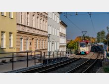 Straßenbahnstation in einer städtischen Umgebung mit einer beige-roten Straßenbahn auf einem erhöhten Streckengleis. Links befinden sich mehrere mehrstöckige Wohnhäuser in Pastellfarben, rechts Bäume mit herbstlich gefärbtem Laub.