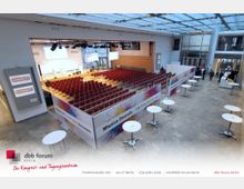 Das Bild zeigt einen modernen Tagungsraum im dbb forum Berlin mit einer Bestuhlung aus rot gepolsterten Stühlen, ausgerichtet auf eine Bühne mit Podium und Leinwänden. Im Vordergrund stehen Stehtische, und die Umgebung ist von einer offenen Glasarchitektur geprägt.