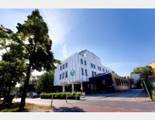 Modernes, mehrstöckiges Gebäude mit weißer Fassade und großen Fenstern, ergänzt durch einen schwarz verkleideten Anbau mit der Beschriftung „Café“. Im Vordergrund sind Grünflächen, Bäume und ein Bürgersteig sichtbar, der von Parkplätzen und Fahrrädern gesäumt ist.