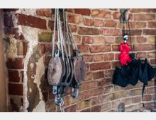 Alte Backsteinwand mit sichtbarem Verputzschaden, an der ein hölzerner Flaschenzug mit Seilen sowie mehrere gefaltete Regenschirme, darunter ein roter und ein schwarzer, aufgehängt sind.