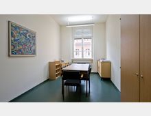 Ein schlicht eingerichteter Büroraum mit einem Tisch und vier Stühlen in der Mitte. An der linken Wand hängt ein abstraktes Gemälde, gegenüber steht ein Schrank, und ein großes Fenster mit weißem Rahmen lässt Tageslicht herein.