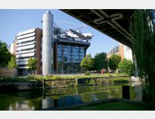 Moderne Architektur des Deutschen Technikmuseums in Berlin mit einer großen Glasfassade und einem über dem Gebäude angebrachten Flugzeug. Im Vordergrund ein Kanal mit bewachsenen Ufern, umgeben von Bäumen und städtischen Gebäuden.