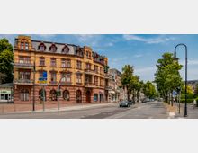 Straßenansicht mit einem historischen, gelb-braunen Backsteingebäude mit mehreren Etagen, Mansarddach und Balkonen, das eine Apotheke beherbergt. Neben dem Gebäude verläuft eine belebte Straße mit Autos, Bäumen und Straßenlaternen auf beiden Seiten.