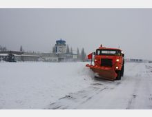 Ein orangefarbener Schneepflug räumt eine verschneite Landebahn, im Hintergrund ist ein Flughafengebäude mit einem Kontrollturm und umliegenden Gebäuden sichtbar. Der Himmel ist grau und die Umgebung von Schnee bedeckt.
