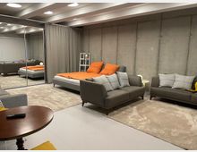 Industrial-style bedroom-living space with exposed concrete walls, a low bed with orange bedding, and gray sofas on large area rugs. Mirrored sliding closet doors and a ceiling with recessed circular lights frame the room, with a curtain divider and small side tables along the wall.