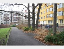 Gehweg in einer städtischen Parkanlage mit kahlen Bäumen und Sträuchern im Herbst, flankiert von einem grünen Rasen auf der linken Seite und einem Zaun vor mehrstöckigen Wohngebäuden auf der rechten Seite.
