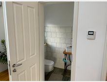 Blick in ein kleines Badezimmer mit weiß gefliesten Wänden, Keramik-Waschbecken auf einem Holzgestell und einer hängenden Toilette. Im Vordergrund ist eine geöffnete weiße Tür zu sehen, daneben hängt ein Wandthermostat an der Wand.