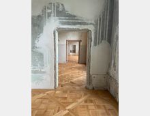 Blick durch mehrere Türöffnungen in eine Reihe leerer Innenräume mit neu verlegtem Holzparkett im geometrischen Muster. Die Wände sind teilweise roh und mit ungleichmäßigem Putz sowie sichtbaren Renovierungsspuren versehen.