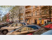 Straßenszene mit mehreren parkenden Autos im Vordergrund und hintereinander gereihten Altbau-Wohnhäusern im Hintergrund, teils mit Balkonen und verzierten Fassaden. Neben einem der Gebäude befindet sich ein Geschäft mit kyrillischer Beschriftung und Speisekarten im Schaufenster.