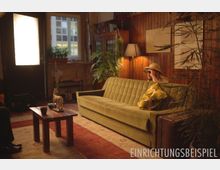Wohnzimmer im Retro-Stil mit grüner Polstercouch, Holzwänden und warmem Licht; im Vordergrund ein kleiner Holztisch mit Dekofigur, im Hintergrund Pflanzen, ein Holzregal und ein Fenster. Rechts sitzt eine Person mit Fernglas und Hut auf der Couch.