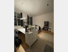 Wohnraum mit hohen Decken und großen Fenstern, durch Vorhänge abgedunkelt. Im Vordergrund steht eine weiße Kommode mit Alltagsgegenständen, dahinter ein Esstisch. Im Hintergrund befinden sich Bücherregale, eine Gitarre und ein Teppich, kombiniert mit einem Holzboden und gefliesten Bereich.