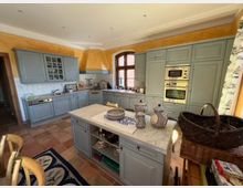 Geräumige Küche im Landhausstil mit blauen Einbauschränken, einer zentralen Kücheninsel mit Marmorarbeitsplatte und Terrakotta-Fliesenboden. Im Hintergrund befindet sich ein großes Fenster mit Holzbogenrahmen, und an den Wänden sind gelbe Akzente sichtbar.