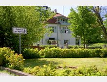 Zweistöckiges Gebäude mit weißer Fassade und roten Dachziegeln, teilweise verdeckt durch Bäume und Sträucher in einem gepflegten Garten. Im Vordergrund steht ein Schild mit der Aufschrift „Ruderklub am Wannsee“.
