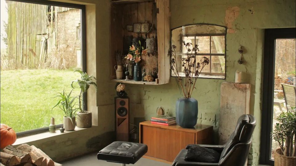 Gemütlicher Wohnraum mit rustikaler Einrichtung, einschließlich grüner Wände, einem großen Fenster mit Blick auf eine Wiese, einem Ledersessel mit Fußhocker und Holzdekor. Neben einer Vitrine befinden sich eine Vase mit getrockneten Blumen und ein Holzschrank mit Büchern und Dekorationsgegenständen.