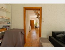 Blick durch einen holzgerahmten Türdurchgang in einen schmalen Flur mit Holzfußboden und weiß strukturierten Wänden. Links stehen Regale, am Ende führt der Flur in einen hellen Raum mit Tisch, Stuhl und Fenster.