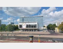 Modernes Gebäudeensemble in einer städtischen Umgebung, bestehend aus einem rechteckigen, niedrigen Bau mit Glasfront und dem Schriftzug „Kino International“ sowie einem hohen, dahinterliegenden Wohn- oder Bürogebäude. Im Vordergrund befinden sich eine mehrspurige Straßenkreuzung, Radfahrer und Fußwege, umgeben von Bäumen und weiteren kleineren Gebäuden.