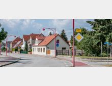 Straßenszene in einem Wohngebiet mit Einfamilienhäusern, teilweise mit roten Ziegeldächern. Die Straße ist von Gehwegen, roten Straßenlaternen und Verkehrsschildern begrenzt; im Hintergrund sind Bäume und blauer Himmel sichtbar.