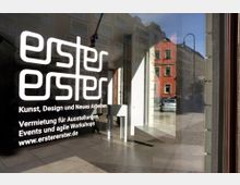 Fensterscheibe eines modernen Ausstellungsraums mit der Aufschrift „erstererster – Kunst, Design und Neues Arbeiten“, umgeben von weiteren Informationen zur Vermietung für Events und Workshops. Im Hintergrund spiegelt sich eine Straßenansicht mit mehrstöckigen Wohnhäusern und parkenden Autos.