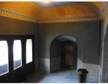 Ein Innenraum mit einer gewölbten Decke und gelben Verzierungsmustern nahe der Deckenränder, einer Tür mit einem Rundbogen und einer verglasten Eingangsfront auf der linken Seite. Die Wände sind in dunklen Farbtönen gestrichen, und der untere Teil ist mit hellen Kacheln versehen.