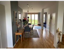 Ein modernes Wohnzimmer mit Holzboden, einem Essbereich mit einem Holztisch und blauen Stühlen sowie einem Wohnzimmerbereich mit einem blauen Sofa. Große Fenster mit Blick auf einen Garten sorgen für viel Tageslicht, und dekorative Elemente wie Pflanzen und Bilder schmücken den Raum.