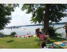 Menschen entspannen auf einer Wiese am Ufer eines Sees, einige liegen auf Decken, andere sitzen auf Stühlen im Schatten eines großen Baumes. Im Hintergrund sind Badende im Wasser und eine kleine Anlegestelle mit Booten zu sehen, umgeben von bewaldeten Ufern.