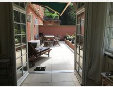 Blick durch eine geöffnete Glas-Doppeltür auf eine Terrasse mit hellen Fliesen, Holzmöbeln und Pflanzenbeeten an den Seiten. Im Hintergrund ist eine rosafarbene Hauswand mit einigen Fenstern sichtbar, darüber Bäume und Grünfläche.