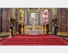 Innenraum einer prunkvollen Kirche mit einem reich verzierten Altar. Der Altarbereich ist mit roten Teppichen bedeckt, flankiert von hohen goldenen Leuchtern, während drei große, bunte Glasfenster biblische Szenen darstellen.