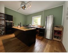 Moderne Küche mit grün gestrichenen Wänden, einem freistehenden Küchenblock mit Holzoberfläche, schwarzen Unterschränken und einem eingebauten Ofen. Ein großer Edelstahlkühlschrank steht rechts, daneben Regale mit Getränken und Lebensmitteln; ein Fenster mit Blick ins Grüne dominiert die Rückwand.