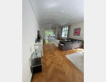 Wohnzimmer mit hellem Fischgrätenparkett, einer grauen Eckcouch und großen Fenstern, die viel Tageslicht hereinlassen. Im Hintergrund ein Essbereich mit Blick auf einen begrünten Außenbereich, ergänzt durch eine farbenfrohe Hängelampe und moderne Dekorationen.