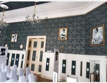 Historischer Raum mit grüner, gemusterter Tapete, verziert mit goldgerahmten Schwarz-Weiß-Fotografien an den Wänden. Ein weiß gestrichener Türrahmen und Holzvertäfelungen mit grünen Einsätzen dominieren die Architektur; weiße Stühle und Ausstellungsdisplays sind ebenfalls zu sehen.