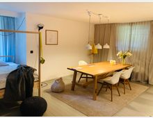 Gemütliches Zimmer mit Holzboden, bestehend aus einem Essbereich mit einem Holztisch, weißen Stühlen und einer Blumen-Dekoration. Daneben befindet sich ein Schlafbereich mit einem Holzbett, getrennt durch einen Vorhang; der Raum ist in warmen Tönen gehalten.