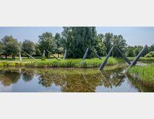 Ein Park mit einem flachen Teich im Vordergrund, umgeben von Schilf und Bäumen. Aus dem Wasser und der angrenzenden Wiese ragen mehrere geneigte, metallische Skulpturen in diagonaler Ausrichtung heraus.