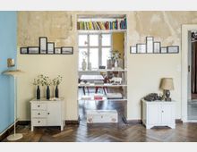 Wohnzimmer mit sichtbaren Gebrauchsspuren an einer beige-blauen Wand; zentral eine offene Regalwand, die einen Blick in ein weiteres Zimmer mit großen Fenstern und einem Esstisch bietet. Der Raum enthält Holzböden, zwei weiße Kommoden mit Dekoration und eine helle Stehlampe.