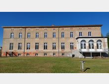Historisches dreigeschossiges Gebäude mit schlichten Fassaden, weißen Sprossenfenstern und einem Anbau auf der rechten Seite mit großen Rundbogenfenstern und einer Terrasse. Das Gebäude steht in einer gepflegten Rasenfläche unter klarem, blauem Himmel.