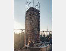 Eine Frau liegt in einer freistehenden Badewanne auf einer Dachterrasse mit Blick auf eine Ziegelmauer und ein schmiedeeisernes Geländer im Sonnenuntergang; im Hintergrund ist die Berliner Fernsehturm-Silhouette sichtbar.