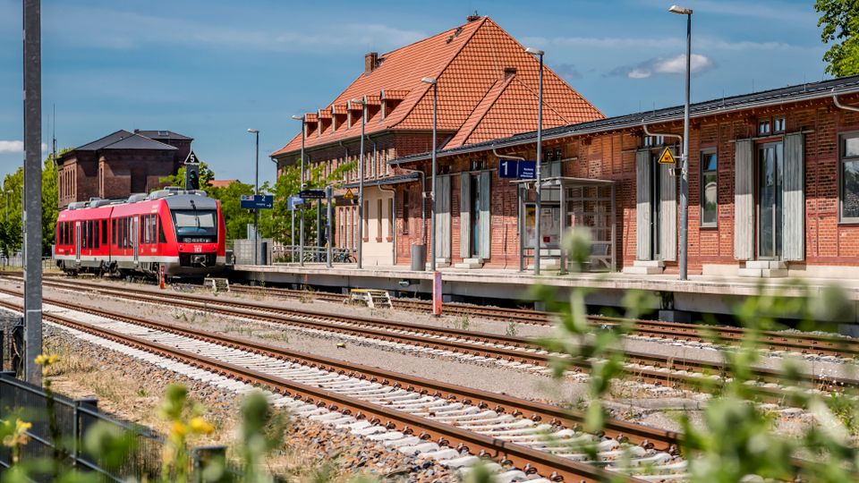 Bahnhof mit mehreren Gleisen und einem Bahnsteig vor einem langen Backsteingebäude mit rotem Ziegeldach, Lampenmasten und Beschilderung. Links steht ein roter Regionalzug an den Gleisen, umgeben von Grünflächen und Bäumen.