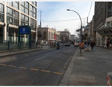 Straßenszene in einer Berliner Innenstadt mit Blick auf die U-Bahn-Station "Oranienburger Tor", flankiert von modernen und historischen Gebäuden. Verkehrssituation mit Autos, einem Roller und Fußgängern, sowie Straßenlaternen, Baustellenabsperrungen und Straßenschildern im Hintergrund.