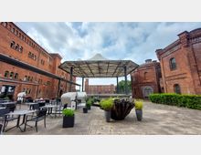 Außenbereich eines historischen Backsteinbaus mit großer Überdachung in der Mitte. Runde Tische und Stühle stehen auf einem gepflasterten Platz, umgeben von Pflanzenkübeln; im Hintergrund weitere Backsteingebäude und ein bewölkter Himmel.