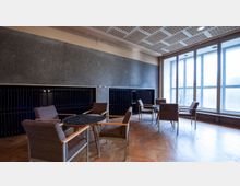 Ein schlicht eingerichteter Raum mit grauen Wänden, hohen Fenstern und dunklen Heizkörpern. Es stehen mehrere kleine Tische mit braunen Stühlen auf einem Parkettboden verteilt.