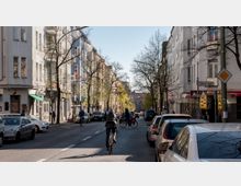 Straßenszene in einer Stadt mit mehrstöckigen Wohngebäuden auf beiden Seiten, parkenden Autos am Straßenrand und Menschen, die Fahrräder fahren oder zu Fuß unterwegs sind. Einige Bäume säumen die Straße, deren Laub beginnt auszutreiben, und im Hintergrund ist eine Ampel sichtbar.