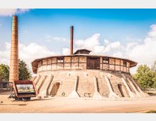 Rundbau eines historischen Ringofens aus Backstein mit sichtbaren Stützstrukturen und einem hohen Schornstein, umgeben von einer offenen Fläche und Bäumen im Hintergrund. Im Vordergrund steht ein alter Förderwagen mit einem Schild, das „Alter Hafen“ zeigt.
