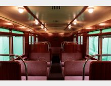 Innenansicht eines historischen Zugwaggons mit gepolsterten Sitzbänken, hölzernen Wandverkleidungen und grün abgedeckten Fenstern. Die Decke ist hell und mit gleichmäßig angeordneten Lampen ausgestattet.