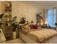 Gemütliches Schlafzimmer mit einem großen Bett, dekoriert mit Kissen und Decken in Erdtönen. Das Zimmer ist mit mehreren Pflanzen, weißen Vorhängen vor einem Fenster sowie Regalen mit Büchern und Dekorationsgegenständen eingerichtet.