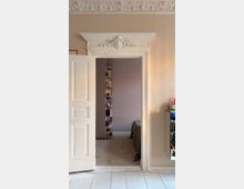 Interior view of a room with a white paneled door open into a bedroom, framed by an ornate white doorway with decorative molding and a patterned plaster ceiling border. Inside, a narrow vertical bookshelf stands against a pale wall beside a bed, with white plank flooring in the foreground.