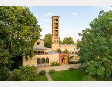 Historische Kirche mit einem hohen Glockenturm im romanischen Stil, umgeben von dichtem Grün und einem gepflegten Garten. Das Gebäude hat Rundbögen, ein rotes Ziegelportal und helle Wände mit einem Mix aus Stein- und Glasdächern.