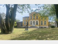 Gelbes zweistöckiges Gebäude im historischen Stil mit weißen Fensterrahmen und einem Eingangsportal mit drei Rundbögen, davor eine Statue auf einer Rasenfläche. Im Vordergrund stehen große Bäume, und der Himmel ist teils bewölkt.