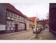 Gepflasterte Straße mit Fachwerkhäusern und weiteren historischen Gebäuden, deren Fassaden in verschiedenen Farben gestrichen sind. Am Straßenrand parken mehrere Autos, und im Hintergrund sind kahle Bäume erkennbar.