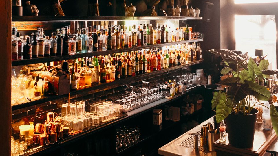 Innenansicht einer Bar mit langen, beleuchteten Regalen voller Spirituosenflaschen und gestapelten Gläsern vor einer dunklen Wand. Rechts steht ein Tresen mit Metallarbeitsfläche neben einem großen Fenster und einer Topfpflanze.